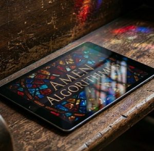 Ein Tablet liegt schräg auf einer dunklen, rustikalen Holzkirchenbank. Auf dem Bildschirm des Tablets ist ein buntes Design zu sehen, das an ein klassisches Kirchen-Buntglasfenster erinnert. Darauf steht in goldenen Großbuchstaben der Text „AMEN ALGORITHMUS“. Farbige Lichtreflexe spiegeln sich auf dem Holz der Bank.